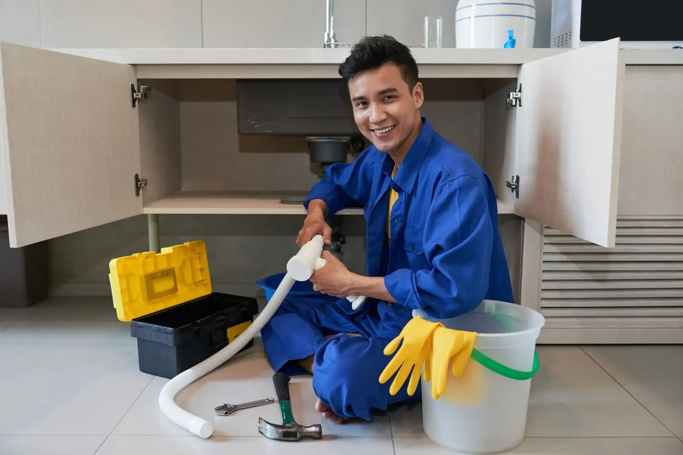 Happy Asian plumber seated on the floor, repairing a kitchen sink.
